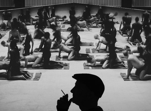 El artista Santiago Sierra, en silueta con gorra y cigarrillo, ante una imagen de su obra Los penetrados