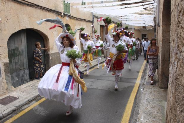 Cossier vestida de blanco con ramos y cintas de colores bailando por una calle de Algaida; al fondo, otros cossiers y una mujer en el portal
