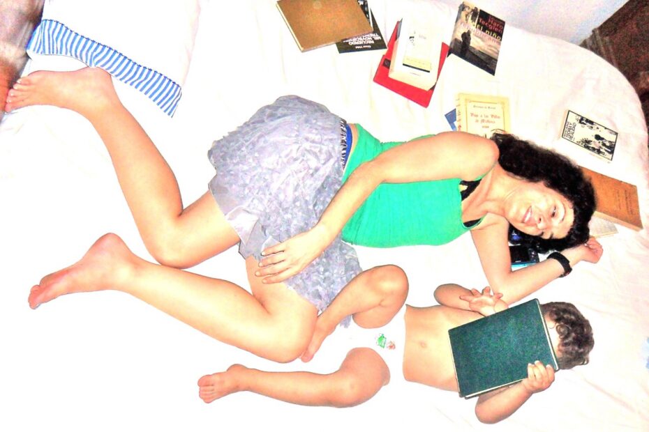 Roser tumbada en una cama blanca rodeada de libros, junto a un niño pequeño que se tapa la cara con un libro verde
