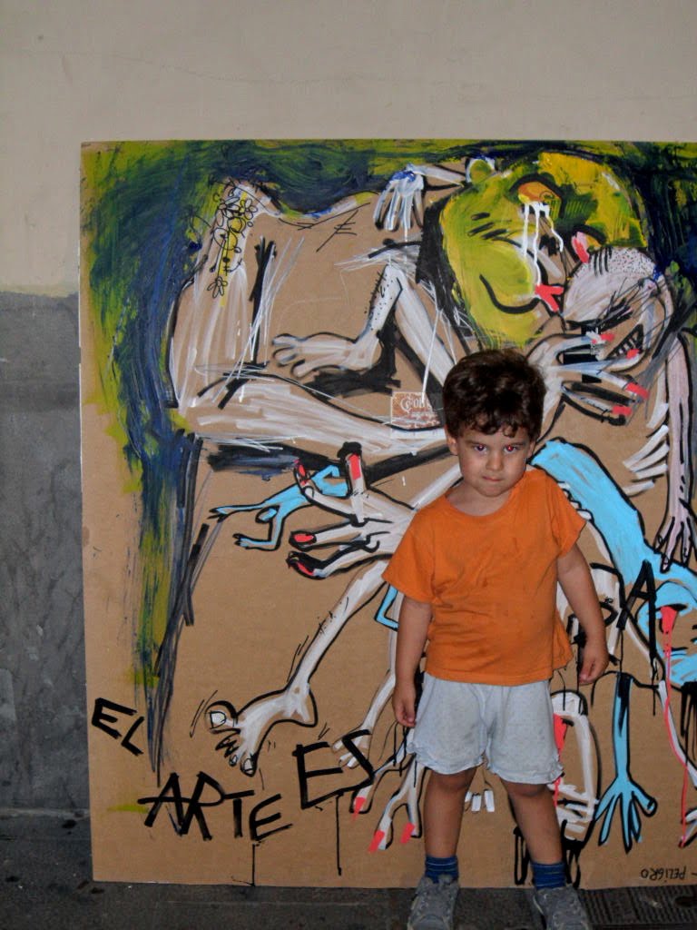 Un niño con camiseta naranja posa de pie delante de la obra «El arte es basura» apoyada en una pared del Raval