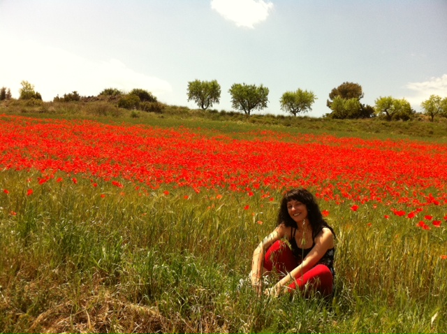 Una mujer de pelo oscuro tumbada en un campo de amapolas rojas, con árboles y un cielo claro al fondo
