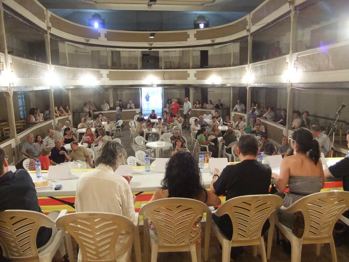 Interior d'un teatre antic amb platea i dos pisos de balconades, amb el públic assegut en cadires de plàstic i una taula amb ponents al davant