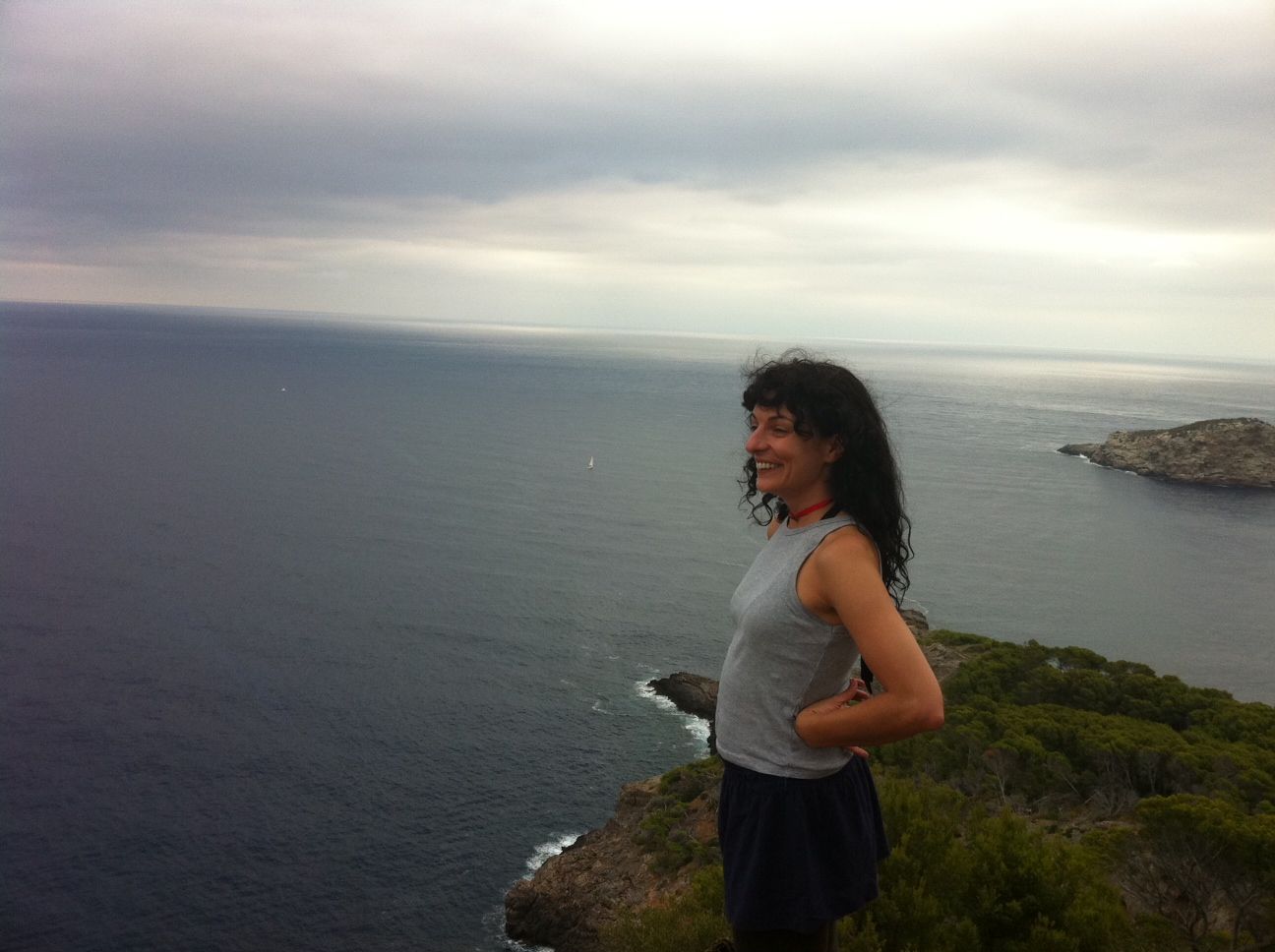 Una dona somrient dalt d'un penya-segat, amb el mar i un illot al fons