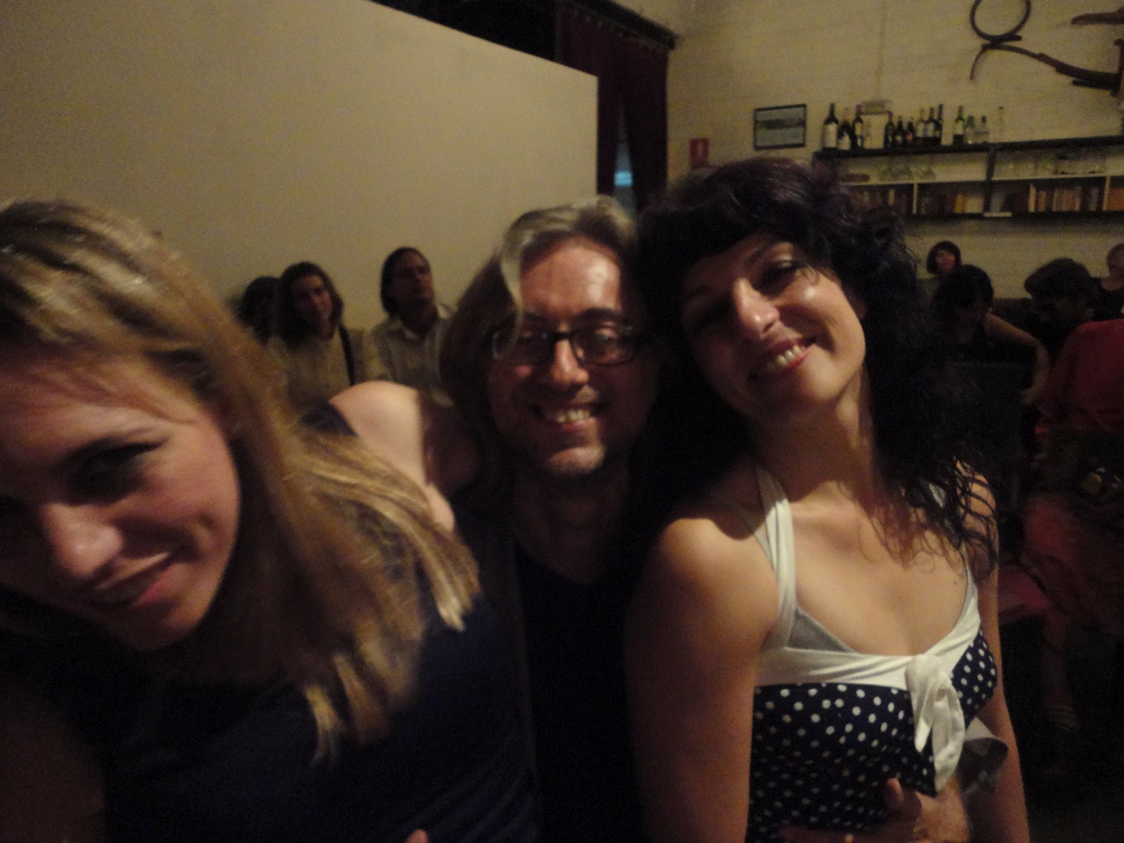 Roser Amills y Víctor Amela sonrientes, junto a una mujer rubia, en un local con público y estantes de botellas al fondo