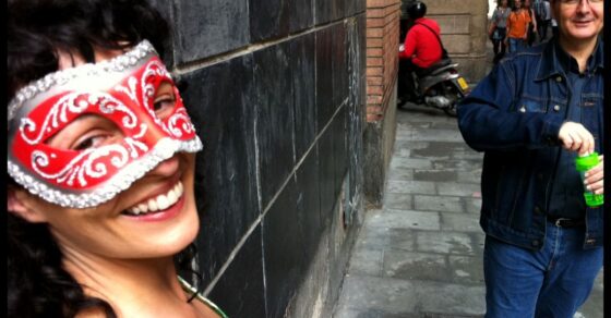 Roser Amills somrient amb un antifaç venecià vermell i blanc, en un carrer de Barcelona; al fons, un home amb jaqueta texana