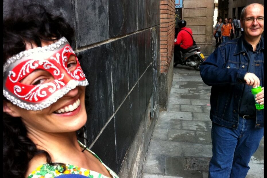 Roser Amills somrient amb un antifaç venecià vermell i blanc, en un carrer de Barcelona; al fons, un home amb jaqueta texana