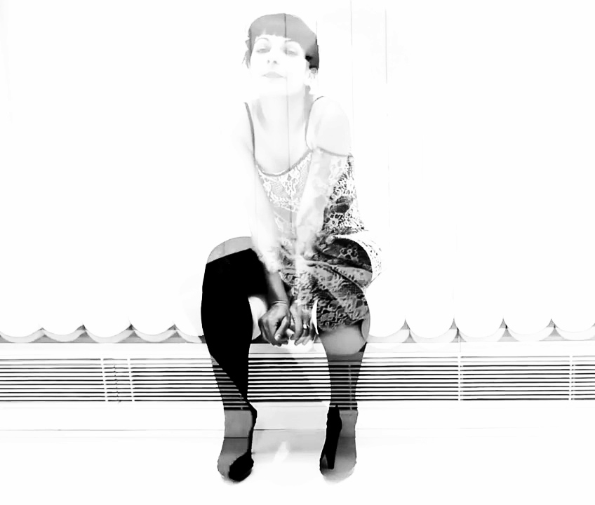 Roser Amills asseguda davant d'una finestra amb persianes, amb mitges negres i vestit estampat, en una fotografia en blanc i negre molt sobreexposada