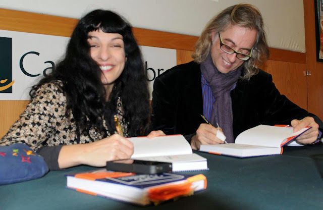 Roser Amills riendo mientras Víctor Amela dedica un ejemplar, con un libro y un móvil sobre la mesa