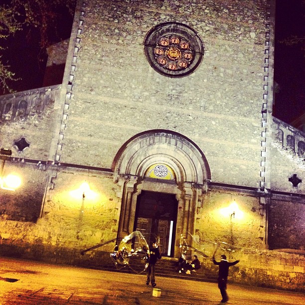 Fachada de una iglesia iluminada de noche, con dos artistas callejeros jugando con aros de pompas en la plaza