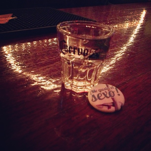 Vaso de chupito de Cuervo sobre una mesa de madera con el pin de «Me gusta el sexo»