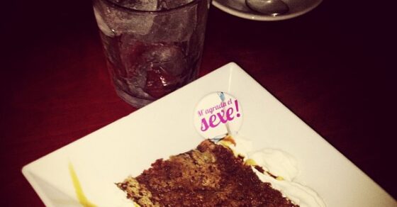 Pin de «M'agrada el sexe» sobre un trozo de tarta de chocolate, junto a un café y un vaso de agua