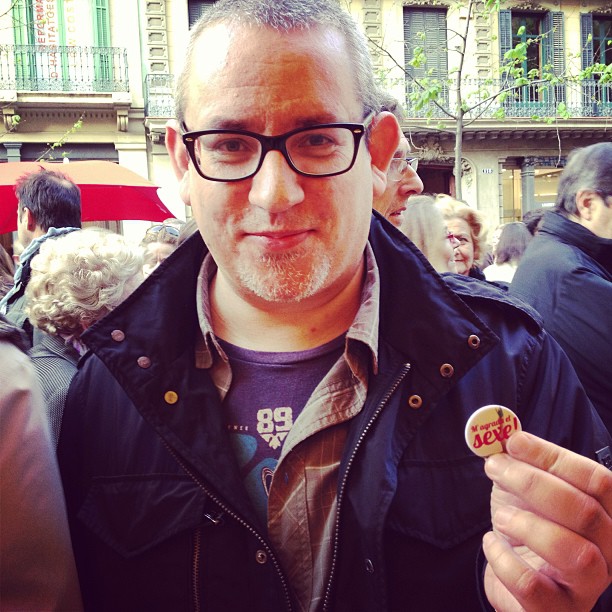 Lector con barba y gafas mostrando el pin de «M'agrada el sexe» en una caseta de Sant Jordi