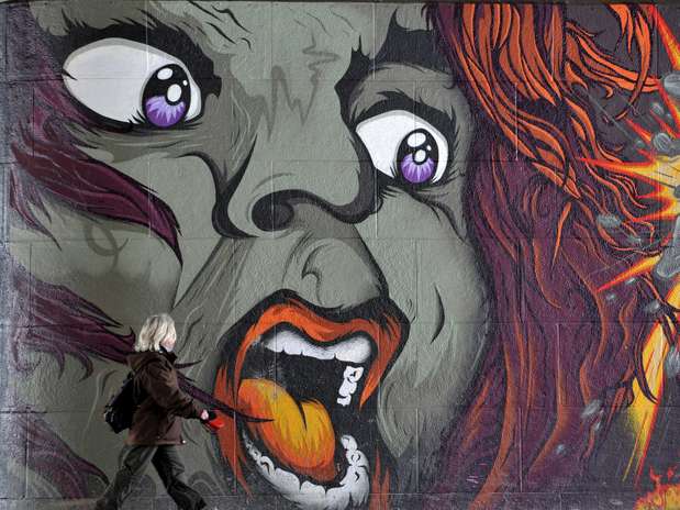 Grafiti de una cara furiosa de ojos morados y boca abierta con una figura diminuta caminando al pie