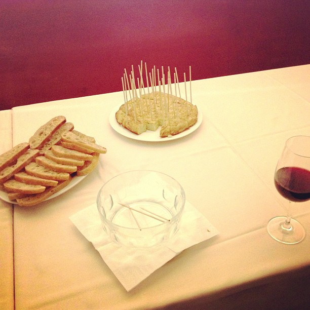 Tortilla de patatas con palillos, pan en rebanadas y una copa de vino tinto sobre un mantel blanco