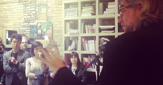 Víctor Amela hablando, de perfil, ante un grupo de asistentes en una sala de paredes de ladrillo