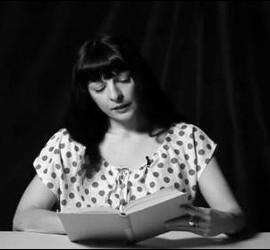 Fotograma en blanco y negro de Roser Amills leyendo un libro ante una mesa, en el vídeo El plaer de la lectura