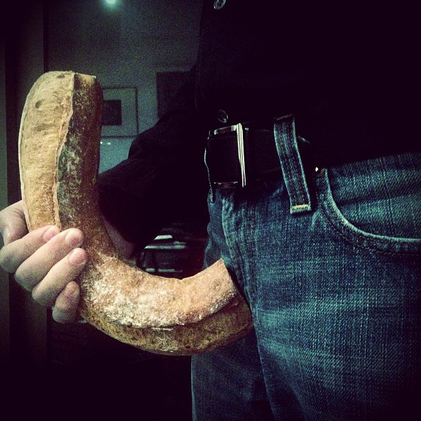 Mano sujetando una barra de pan en forma de herradura, con vaqueros al fondo