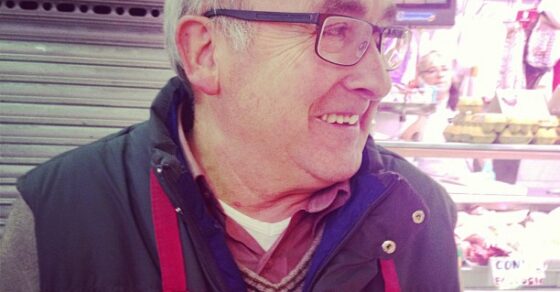 Hombre mayor con gafas, delantal rojo y una chapa de M'agrada el sexe!, ante un puesto del mercado
