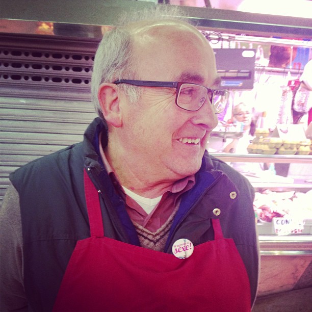Hombre mayor con gafas, delantal rojo y una chapa de M'agrada el sexe!, ante un puesto del mercado