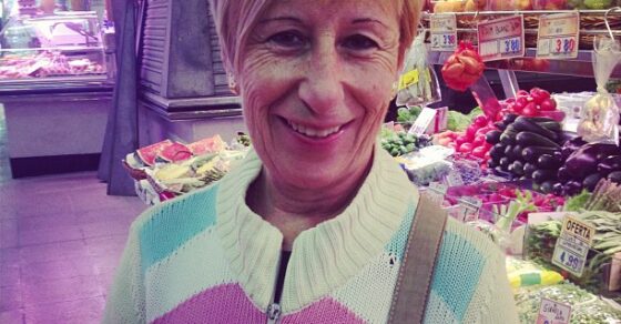 Clienta sonriente con gafas de sol en el pelo y una chapa de ¡Me gusta el sexo!, ante un puesto de fruta del mercado