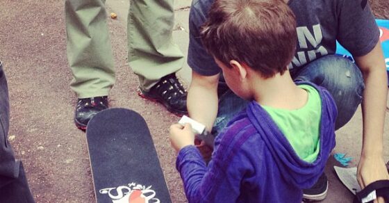 Un chico ayuda a un niño a ponerse las protecciones junto a un monopatín