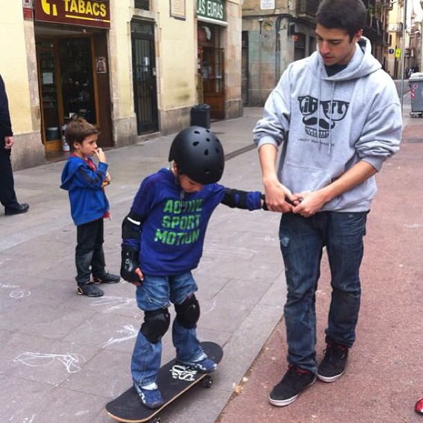Niño con casco y protecciones sobre un monopatín, sujetado por un chico en una calle de Barcelona