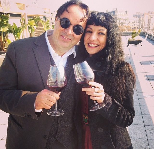 Roser Amills y Oscar Iniesta, con gafas redondas, brindan con copas de vino en una terraza