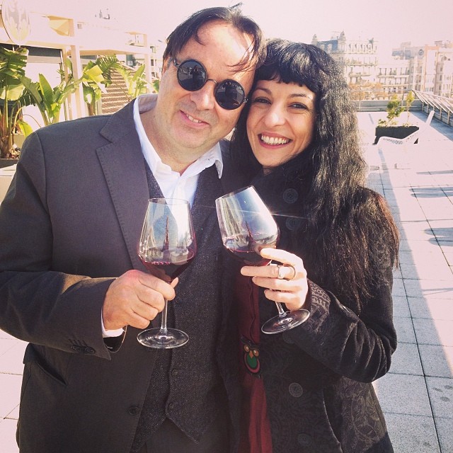 Roser Amills y Oscar Iniesta, con gafas redondas, brindan con copas de vino en una terraza
