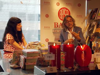 Roser Amills, con vestido de lunares rosas, y Víctor Amela sentados tras una mesa con velas rojas en forma de 110 ante el panel con el logotipo de la librería Bassa