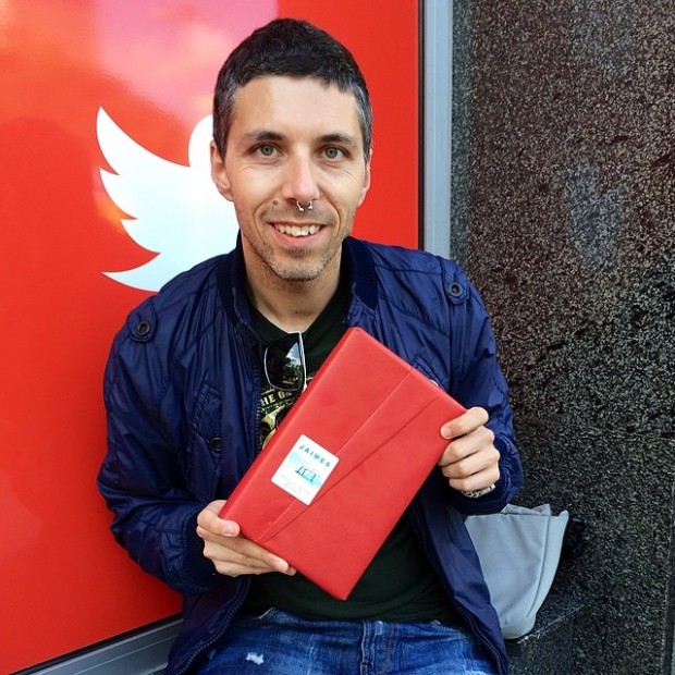 Miquel Requena sonriendo con un libro envuelto en papel rojo ante un cartel de Twitter