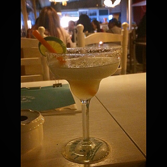 Copa de margarita con el borde escarchado, una rodaja de lima y una pajita naranja, sobre una mesa de madera clara