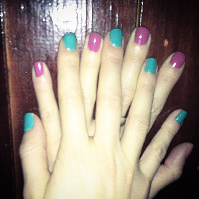 Mano con las uñas pintadas alternando turquesa y fucsia, sobre una superficie de madera oscura