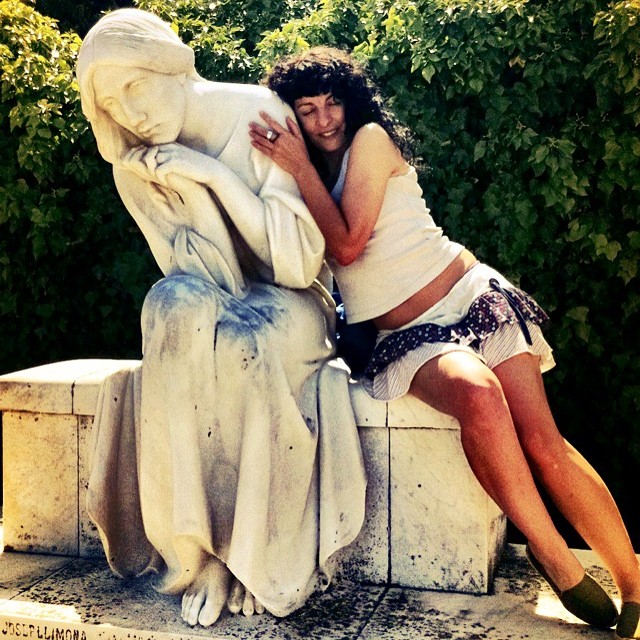 Roser Amills, con el pelo rizado, top blanco y falda estampada, apoyada en el hombro de una escultura blanca de una mujer sentada con la mano en la mejilla