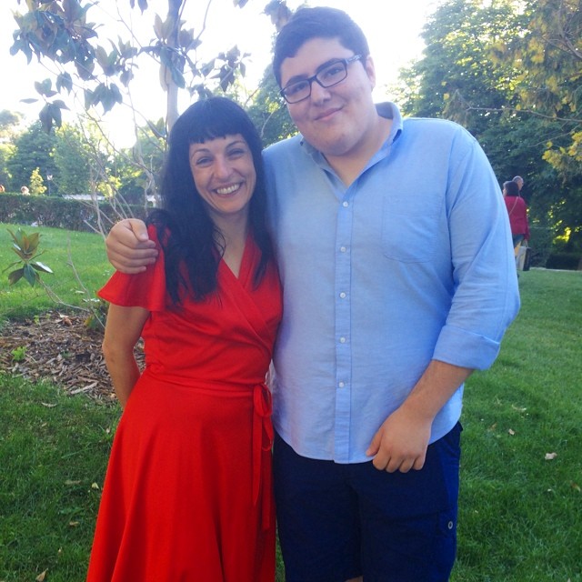 Roser Amills, con vestido rojo, abrazada a un joven con gafas y camisa azul en un parque