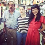 Roser Amills, con vestido rojo, posa sonriendo con dos libreros en una caseta de la Feria del Libro de Madrid