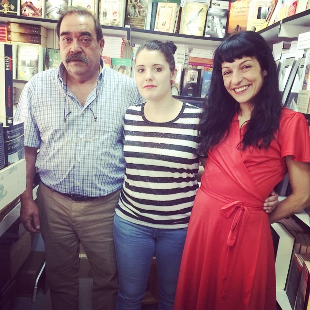 Roser Amills, con vestido rojo, posa sonriendo con dos libreros en una caseta de la Feria del Libro de Madrid