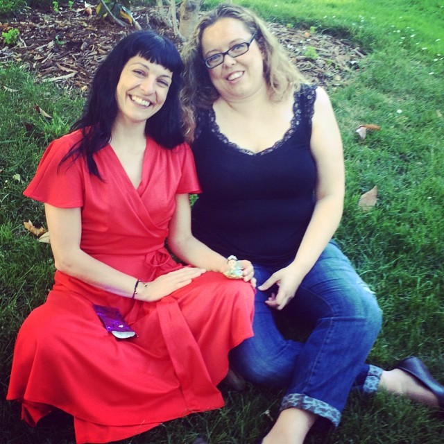 Roser Amills, con vestido rojo, sentada en la hierba junto a una mujer con gafas y blusa negra; entre ambas, una pequeña figura de hada