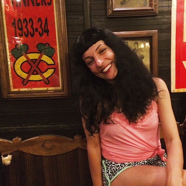 Mujer sonriente con top rosa y pantalón corto de leopardo, ante una pared con carteles antiguos
