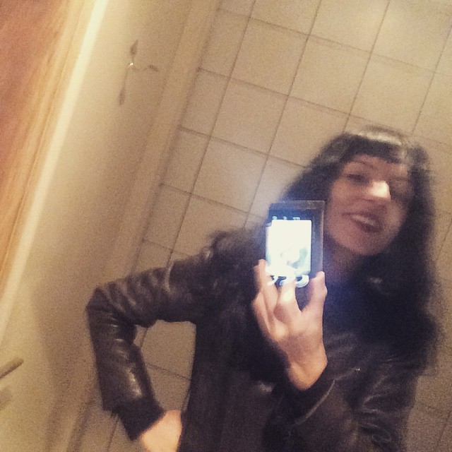 Roser Amills haciéndose un selfi en un espejo con el móvil, con chaqueta de cuero, junto a una pared de azulejos blancos