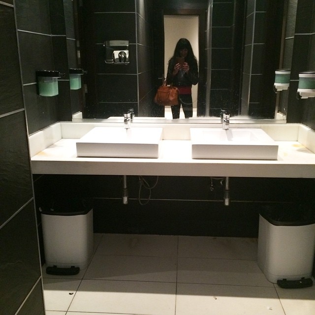 Lavabo doble de un baño público con espejo, en el que se refleja una persona con bolso