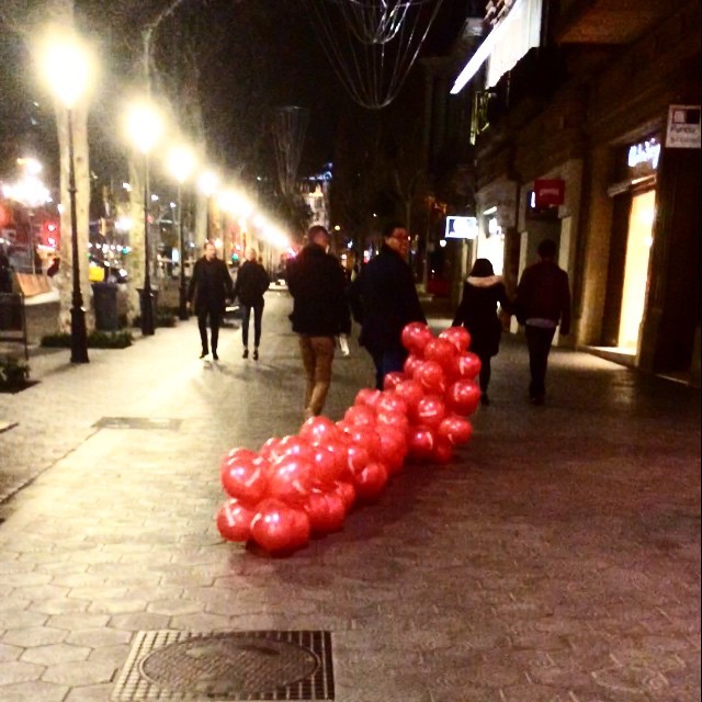 Hilera de globos rojos arrastrada por la acera de noche, con farolas encendidas y varias personas caminando