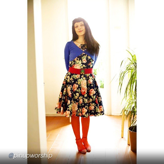 Roser Amills de pie, con rebeca azul, vestido de flores, cinturón rojo y medias rojas, junto a una planta; en la esquina, @pinupworship