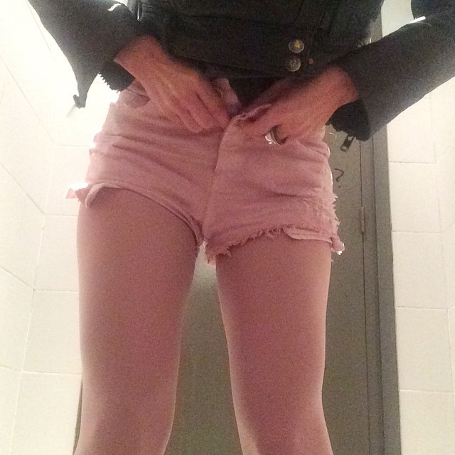 Roser Amills con shorts vaqueros rosas desabrochados en un lavabo de azulejos blancos, serie Amills Public WC