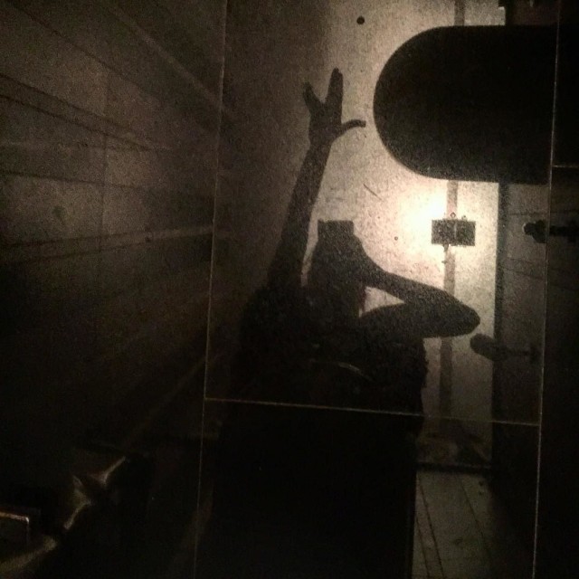 Sombra de una mujer en un lavabo oscuro, con una mano levantada haciendo el saludo vulcano, ante una pared iluminada