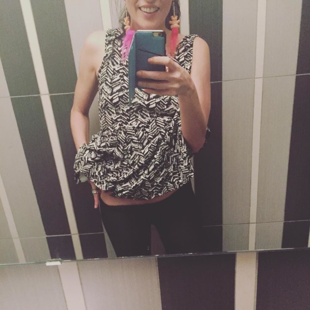 Selfie ante un espejo de rayas verticales: mujer sonriente con blusa estampada en blanco y negro y pendientes de plumas rosas, con el móvil en la mano