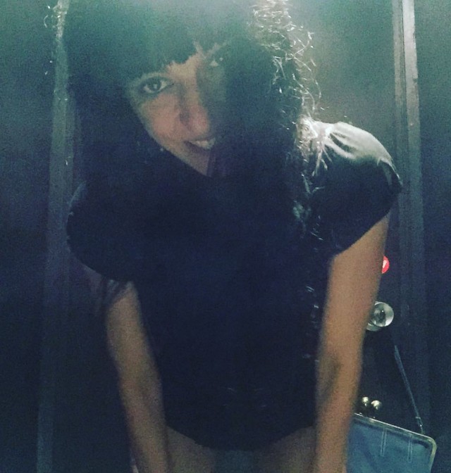 Mujer con flequillo y pelo largo y oscuro, inclinada hacia la cámara y sonriendo, con una camiseta negra, en un lavabo con la puerta al fondo