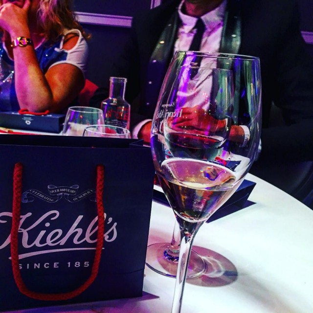 Copa de champán y bolsa de regalo de Kiehl's sobre una mesa redonda, con invitados al fondo