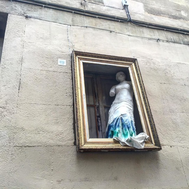 Torso de mujer de yeso, sin brazos y con un paño azul y verde, dentro de un marco dorado colgado en la fachada de un edificio