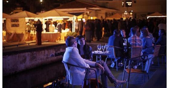 Terraza nocturna junto a un muelle con carpas iluminadas al fondo; en primer plano, una mujer de flequillo conversa con un hombre de chaqueta blanca, con otras personas sentadas a la derecha