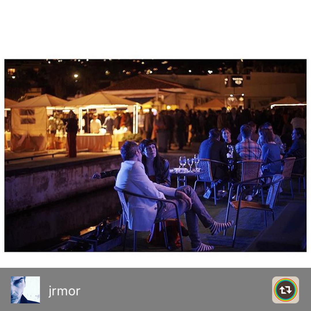 Terraza nocturna junto a un muelle con carpas iluminadas al fondo; en primer plano, una mujer de flequillo conversa con un hombre de chaqueta blanca, con otras personas sentadas a la derecha